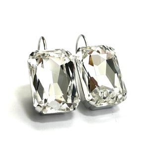 May include: A pair of rectangular, clear crystal earrings with silver-colored hooks. The crystals are faceted, reflecting light and creating a sparkling effect. The earrings are set against a white background.