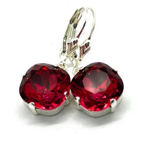 May include: A pair of leverback earrings featuring large, faceted, ruby-red gemstones. The gemstones are set in a silver-colored metal. The earrings are displayed against a white background, highlighting the vibrant color and sparkle of the stones.