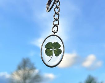 May include: A silver keychain with a four-leaf clover encased in a clear, oval-shaped resin pendant. The clover is a vibrant green against a backdrop of a bright blue sky with fluffy white clouds. The keychain is suspended in the air.
