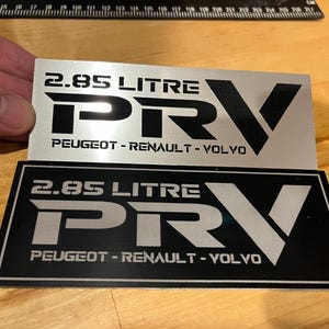 May include: Two silver metal plaques with black lettering. The top plaque reads "2.85 LITRE PRV PEUGEOT-RENAULT-VOLVO". The bottom plaque reads "2.85 LITRE PRV PEUGEOT-RENAULT-VOLVO".