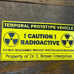 May include: A yellow rectangular sign with black text. The sign reads "TEMPORAL PROTOTYPE VEHICLE," "! CAUTION! RADIOACTIVE," and "DO NOT DISASSEMBLE WITHOUT WRITTEN AUTHORIZATION." It also includes the text "Property of Dr. E. Brown Enterprises" and radiation symbols.