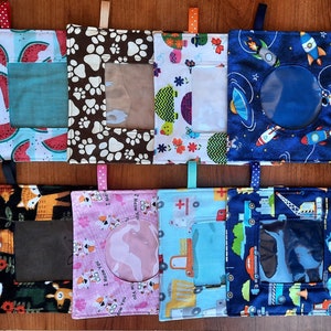 May include: Eight colorful fabric squares with a clear window in the center. Each square features a different pattern, including watermelons, paw prints, flowers, rockets, foxes, cats, construction vehicles, and a cross.