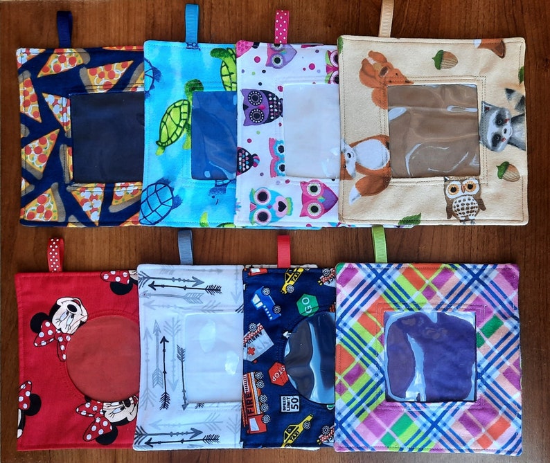 May include: Eight square fabric pouches with hanging loops, each featuring a clear plastic window. Patterns include pizza slices, turtles, colorful owls, woodland animals, cartoon mice, arrows, city traffic signs, and a multi-colored plaid design.