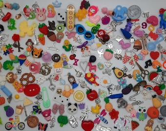 May include: A large collection of assorted colorful charms and miniature figurines including food items, animals, characters, and everyday objects scattered across a white surface, featuring a variety of plastic and metal materials.