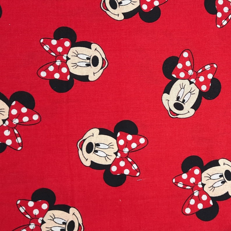 May include: A red fabric featuring a repeating pattern of Minnie Mouse faces. Each character has black ears, a cream-colored face, and a large red bow with white polka dots. The design is printed across the entire surface of the textile material.
