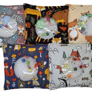 Puede incluir: Cinco bolsas sensoriales cuadradas de tela con varios estampados de animales y naturaleza. Cada bolsa tiene una ventana circular de pl&aacute;stico transparente en el centro, que muestra peque&ntilde;as cuentas de colores y cubos con letras en un relleno granular blanco.