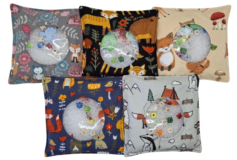 Puede incluir: Cinco bolsas sensoriales cuadradas de tela con varios estampados de animales y naturaleza. Cada bolsa tiene una ventana circular de pl&aacute;stico transparente en el centro, que muestra peque&ntilde;as cuentas de colores y cubos con letras en un relleno granular blanco.