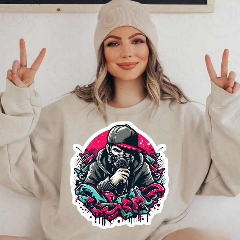 May include: Beige sweatshirt featuring a colorful graffiti-style graphic of a person wearing a gas mask and a baseball cap. The graphic is set against a pink background with red and blue accents. The person in the image is wearing a beige beanie.