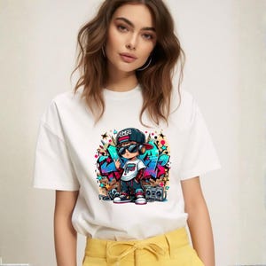 May include: White t-shirt featuring a colorful graphic of a cartoon boy in a hip-hop style outfit, wearing sunglasses and a backwards cap. The design includes graffiti-style art and a boombox. The shirt is worn by a person.
