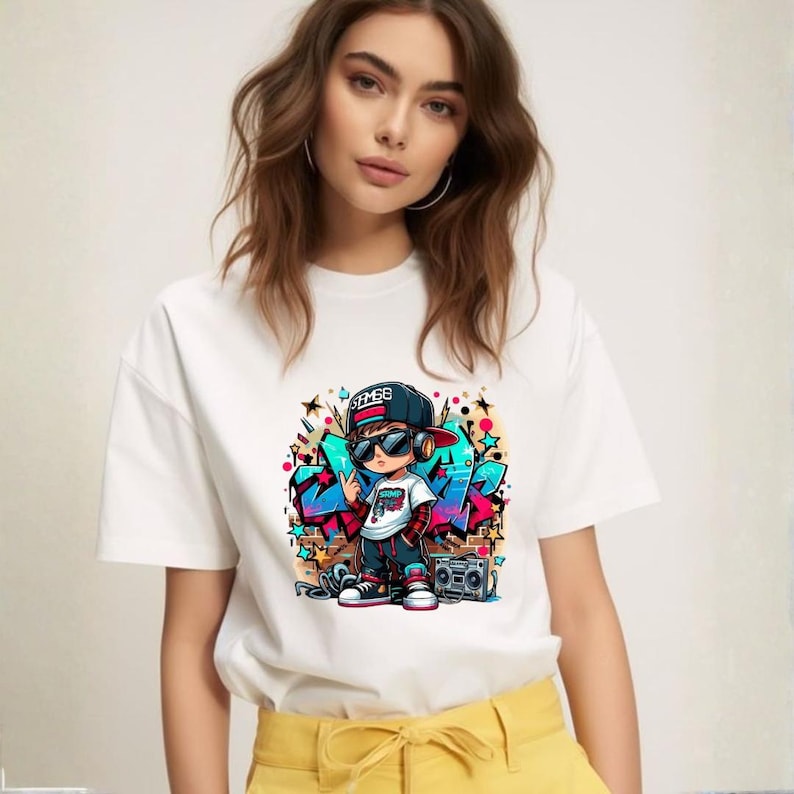 May include: White t-shirt featuring a colorful graphic of a cartoon boy in a hip-hop style outfit, wearing sunglasses and a backwards cap. The design includes graffiti-style art and a boombox. The shirt is worn by a person.