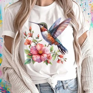 Puede incluir: Camiseta blanca con un colibr&iacute; colorido y flores rosas y naranjas. El ave tiene plumas azules, naranjas y verdes. Un dise&ntilde;o vibrante con tem&aacute;tica de la naturaleza.