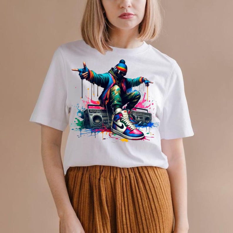 May include: White t-shirt featuring a colorful graphic of a person in a hip-hop style pose, wearing a beanie, sunglasses, and sneakers. The design includes a boombox and paint splatter effects. The person is squatting down with arms outstretched.