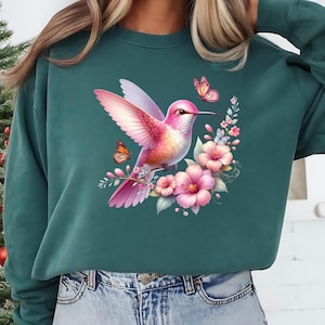 Puede incluir: Sudadera verde azulado con una colorida ilustraci&oacute;n de un colibr&iacute; rosa, flores rosas y mariposas. El dise&ntilde;o est&aacute; centrado en la parte delantera de la sudadera, creando una est&eacute;tica inspirada en la naturaleza. Prenda c&oacute;moda e informal.