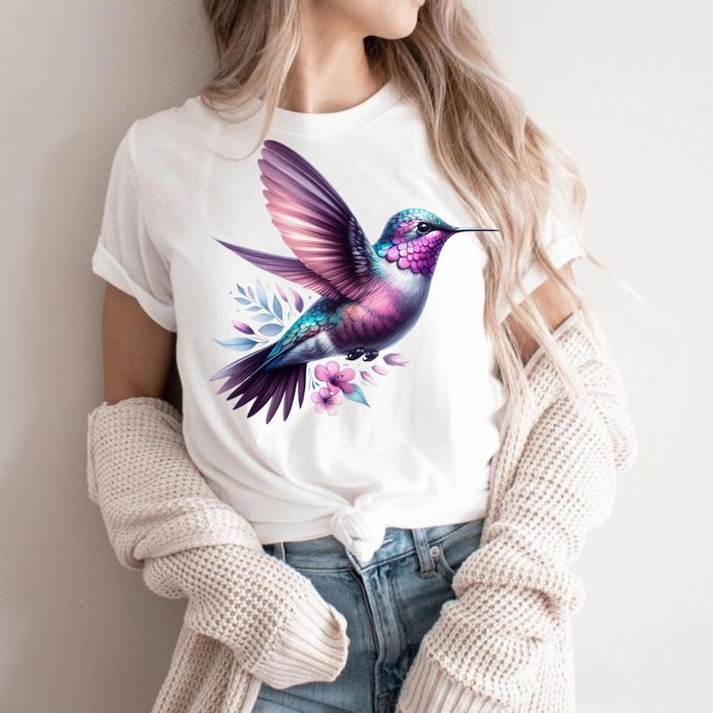 Puede incluir: Camiseta blanca con un dise&ntilde;o de colibr&iacute; en acuarela. El ave presenta plumas moradas, rosas y turquesas, con las alas extendidas. Delicados detalles florales y hojas rodean el colibr&iacute;, creando una est&eacute;tica inspirada en la naturaleza.