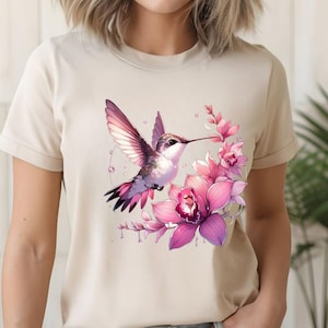 Puede incluir: Una camiseta beige con una ilustraci&oacute;n de acuarela de un colibr&iacute; con alas rosas y moradas, cerca de orqu&iacute;deas rosas. El dise&ntilde;o presenta detalles delicados y una paleta de colores suaves.