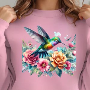 Puede incluir: Sudadera rosa claro con un dise&ntilde;o de colibr&iacute; vibrante con plumaje colorido, flotando cerca de un grupo de flores. El dise&ntilde;o incluye flores rosas, naranjas y azules, con una peque&ntilde;a mariposa. La sudadera es de color rosa claro s&oacute;lido.
