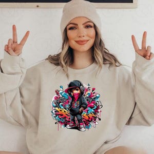 May include: Beige sweatshirt featuring a colorful graphic of a cartoon character in a black hoodie, red bandana, and cap, surrounded by graffiti art. The character is wearing headphones. The sweatshirt is a neutral color.