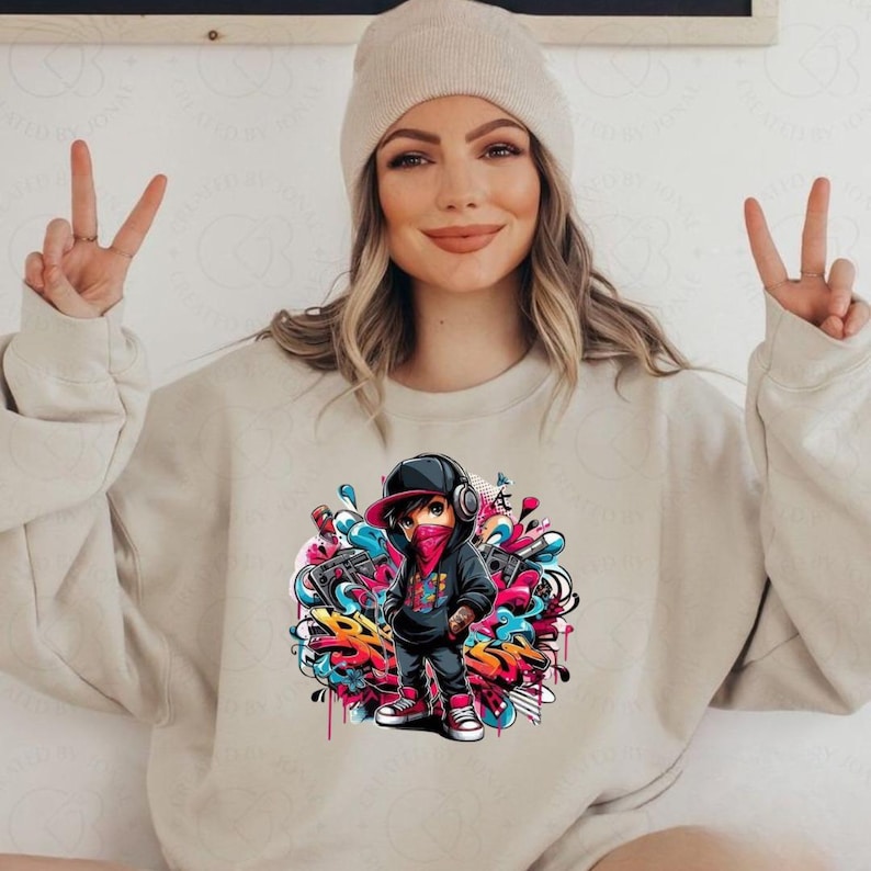 May include: Beige sweatshirt featuring a colorful graphic of a cartoon character in a black hoodie, red bandana, and cap, surrounded by graffiti art. The character is wearing headphones. The sweatshirt is a neutral color.