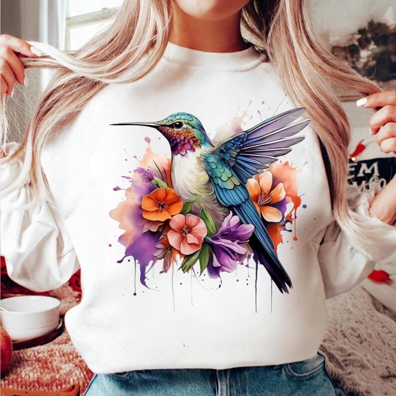 Puede incluir: Sudadera blanca con un dise&ntilde;o de acuarela de un colibr&iacute; con plumas azules, verdes y moradas, rodeado de flores naranjas y moradas. El p&aacute;jaro est&aacute; en vuelo, con las alas extendidas. El dise&ntilde;o tiene un efecto de salpicadura.
