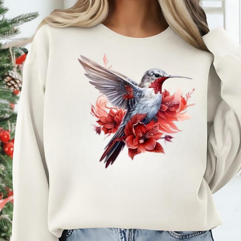 Puede incluir: Sudadera color crema con una ilustraci&oacute;n detallada de un colibr&iacute; con las alas extendidas. El ave tiene plumas grises y blancas con la garganta roja, rodeada de flores rojas vibrantes. El dise&ntilde;o est&aacute; centrado en la parte delantera de la prenda.