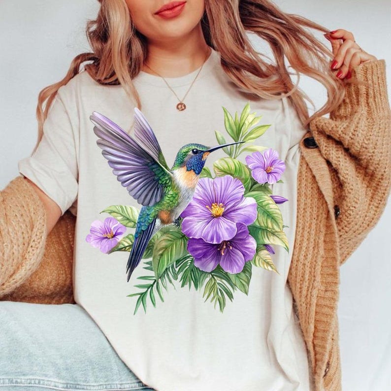 Puede incluir: Camiseta beige con una ilustraci&oacute;n de un colibr&iacute; con las alas extendidas, rodeado de flores moradas y hojas verdes. El p&aacute;jaro tiene plumas azules, verdes y moradas. El dise&ntilde;o est&aacute; centrado en la parte delantera de la camiseta.
