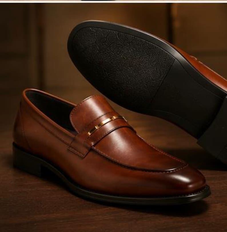 May include: Brown leather loafer shoes with a gold-colored strap detail. The sole of a second shoe is visible in the background. The shoes have a classic design and are likely for formal or business wear.