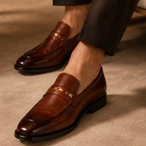 May include: Brown leather loafers with a gold-colored strap across the top. The shoes have a classic design with a rounded toe and a low heel. The image shows the shoes on a person's feet, suggesting a focus on footwear and fashion.