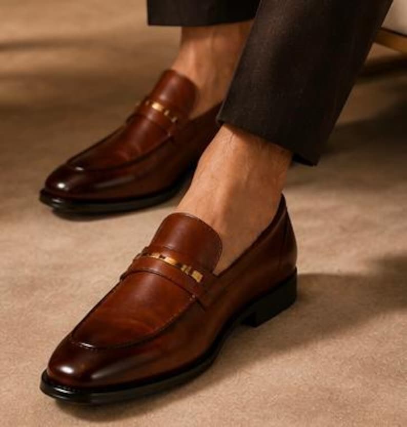 May include: Brown leather loafers with a gold-colored strap across the top. The shoes have a classic design with a rounded toe and a low heel. The image shows the shoes on a person's feet, suggesting a focus on footwear and fashion.