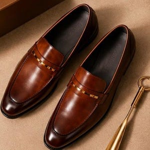May include: A pair of brown leather loafers with a gold-colored accent band. The shoes have a classic design with a polished finish. A gold-colored shoe horn is also visible.