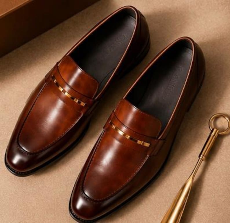May include: A pair of brown leather loafers with a gold-colored accent band. The shoes have a classic design with a polished finish. A gold-colored shoe horn is also visible.