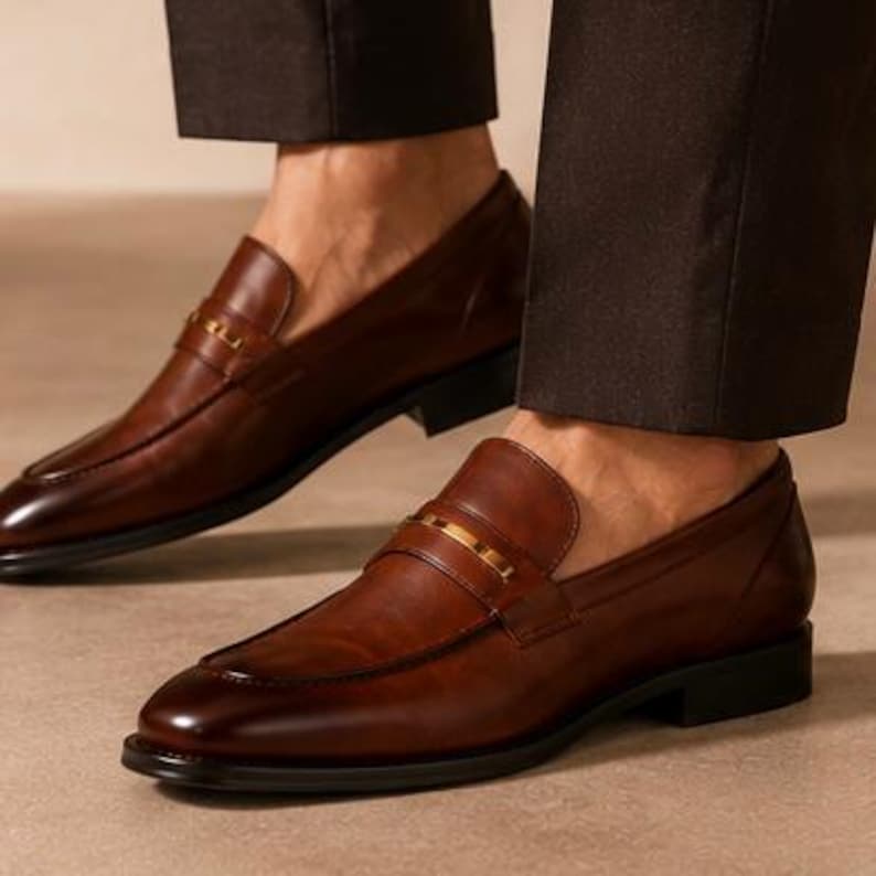 May include: Close-up of two brown leather loafers with a gold-colored strap across the top. The shoes have a classic design with a rounded toe and a low heel. The loafers are worn with brown trousers.