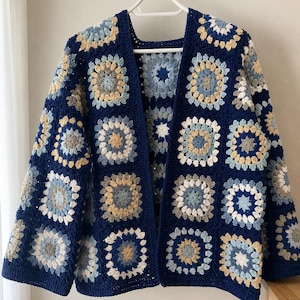 May include: A long-sleeved cardigan made of crocheted granny squares in shades of navy blue, light blue, cream, and grey. The garment features an open front design with a solid navy blue border along the edges and is displayed on a white hanger.