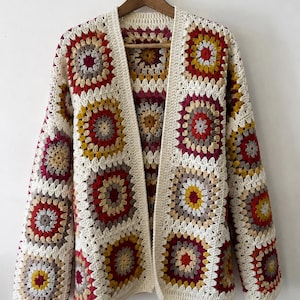 May include: A long-sleeved cardigan made of crocheted granny squares in a repeating pattern of cream, mustard yellow, deep red, and grey. The garment features an open front design with a solid cream-colored crocheted border along the edges.