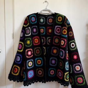 May include: A long-sleeved cardigan made from a black yarn base featuring a repeating pattern of multi-colored granny squares. Each square contains a vibrant circular center in shades of pink, yellow, blue, green, and purple, framed by black.