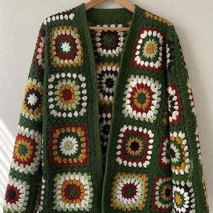 May include: A long-sleeve cardigan made of crocheted granny squares in shades of forest green, mustard yellow, burgundy, and white. The garment features an open front design with a solid dark green border and is displayed on a wooden hanger.