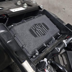 Op de afbeelding: Een zwarte motorfiets opbergbox met een gestructureerd oppervlak en het woord "KSR" erop gegraveerd.