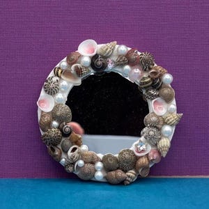 May include: A circular mirror frame decorated with an assortment of small brown, tan, and white seashells interspersed with smooth white pearls. The frame features a variety of spiral and cone-shaped shells, creating a textured border around the glass.