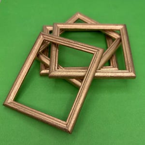 May include: Several small, square, gold-colored picture frames are arranged on a green surface. The frames have a simple, classic design with a slightly textured appearance. The frames are empty, ready to hold photos or artwork.