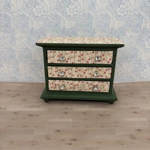 May include: A miniature green chest of drawers with three drawers. The drawer fronts feature a floral pattern with red flowers on a cream background. Each drawer has a crystal knob. The top of the chest also has the floral pattern.