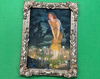 May include: A framed painting of a woman in a yellow dress standing in a forest with glowing lights. The frame is gold and ornate.