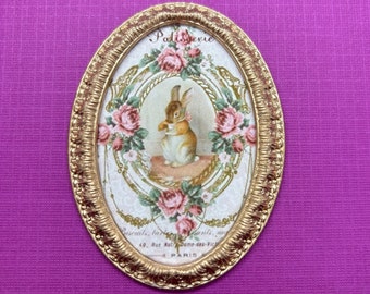 Peut inclure: Un cadre ovale doré avec un fond blanc et une image d'un lapin tenant une fleur. Le cadre a une bordure décorative et le texte "Patisserie Biscuits, la tarte aux fruits, ma 40, Rut Notre-Dame-des-Vic PARIS" est imprimé en bas.