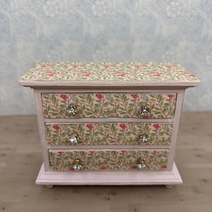 May include: A small wooden chest of drawers painted in a soft pink finish. The top surface and the front of the three drawers feature a decorative pattern of green leaves and small red flowers. Each drawer is fitted with two clear crystal knobs.
