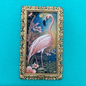 May include: A framed print of a pink flamingo standing in a tropical garden setting. The frame is gold and ornate.