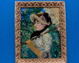 Peut inclure: Une peinture encadrée d'une femme en robe et chapeau blancs, tenant un bâton de marche. L'arrière-plan est un paysage vert et bleu.