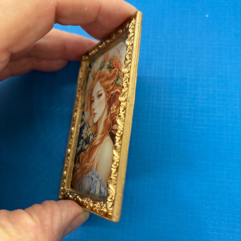 May include: A framed portrait of a woman with long, flowing red hair. She is wearing a floral crown and a blue dress. The frame is gold and ornate.