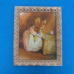 May include: A framed illustration of a brown mouse wearing a pink and white dress, holding a yellow cloth and standing at a table with a white cup and other objects. The frame is gold with an ornate design.