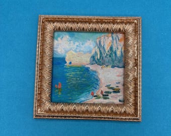May include: A small framed painting of a coastal scene with cliffs, a beach, and the sea. The painting has a gold-coloured ornate frame. The artwork uses a palette of blues, greens, and beige.