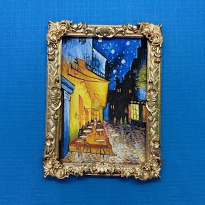 May include: A framed painting of a cafe at night with a starry sky. The painting is in a gold frame with ornate details. The cafe is lit up with yellow lights and there are people sitting at tables outside.