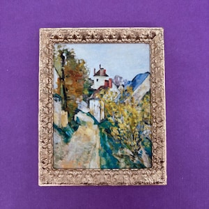 May include: A small framed painting depicting a landscape with buildings, trees, and a road. The painting features a gold-colored ornate frame. The artwork uses a variety of colors, including blue, green, yellow, and brown.