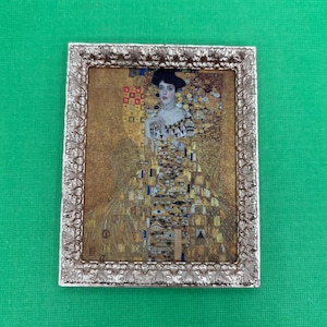 May include: A framed print of a Gustav Klimt painting, featuring a woman in an ornate gold and patterned dress. The frame is silver with a textured design. The artwork has a gold background with geometric shapes.